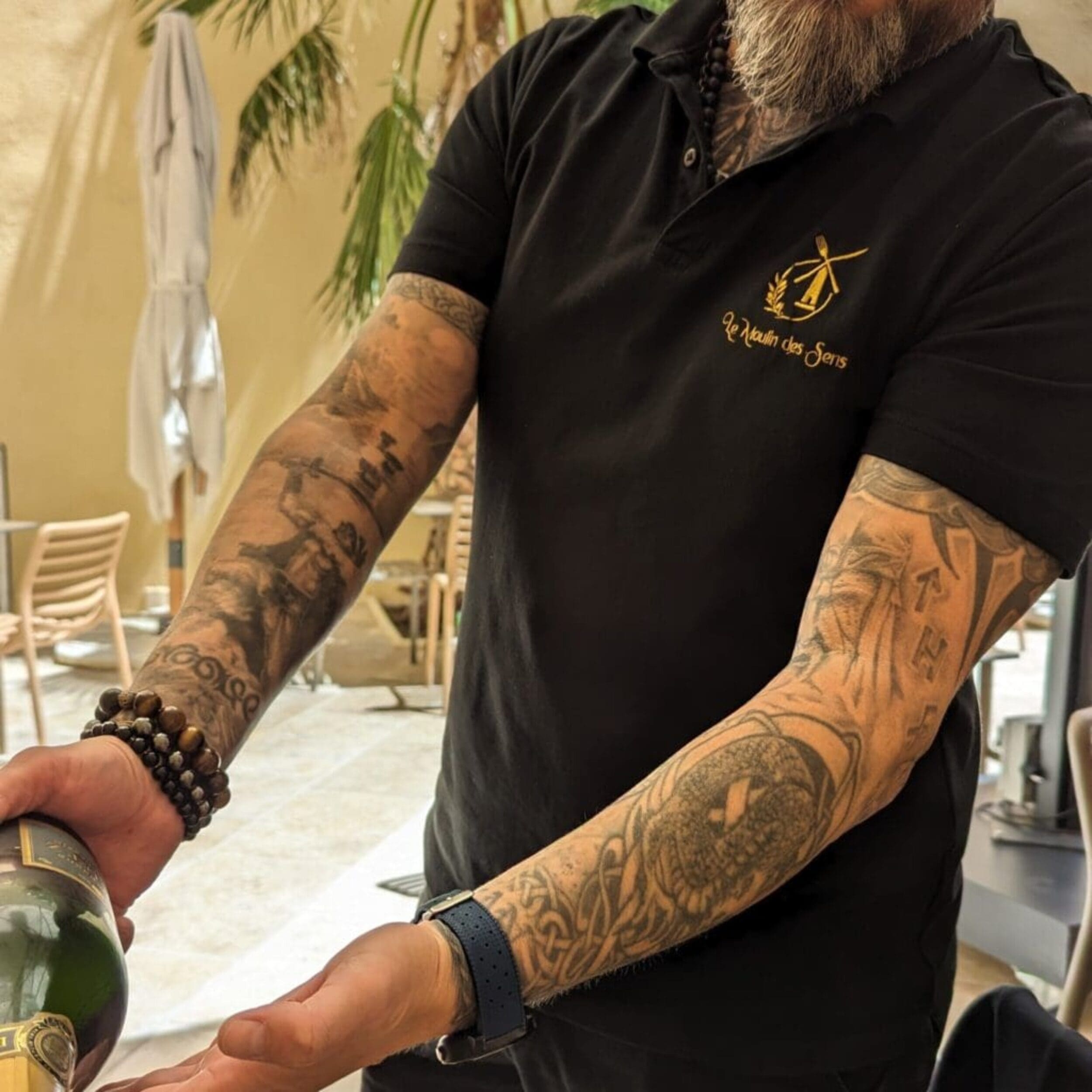Homme servant du champagne, vêtu d’un polo noir avec le logo brodé doré du restaurant Le Moulin des Sens.