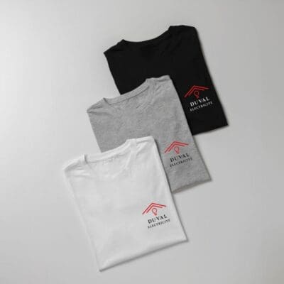 Trois t-shirts pliés (noir, gris et blanc) avec le logo de Duval Électricité imprimé sur la poitrine, représentant un toit stylisé rouge et une ampoule.