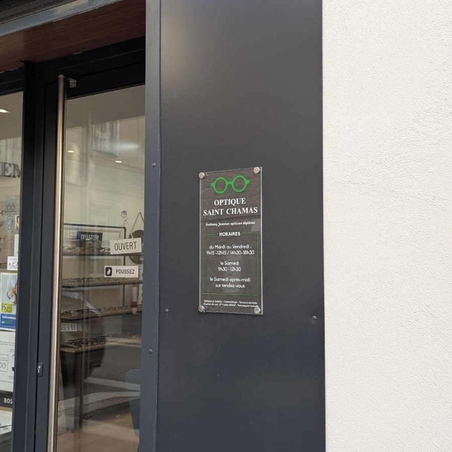 Plaque d'identification "Optique Saint Chamas" fixée à l'entrée d'une boutique, avec horaires d'ouverture.
