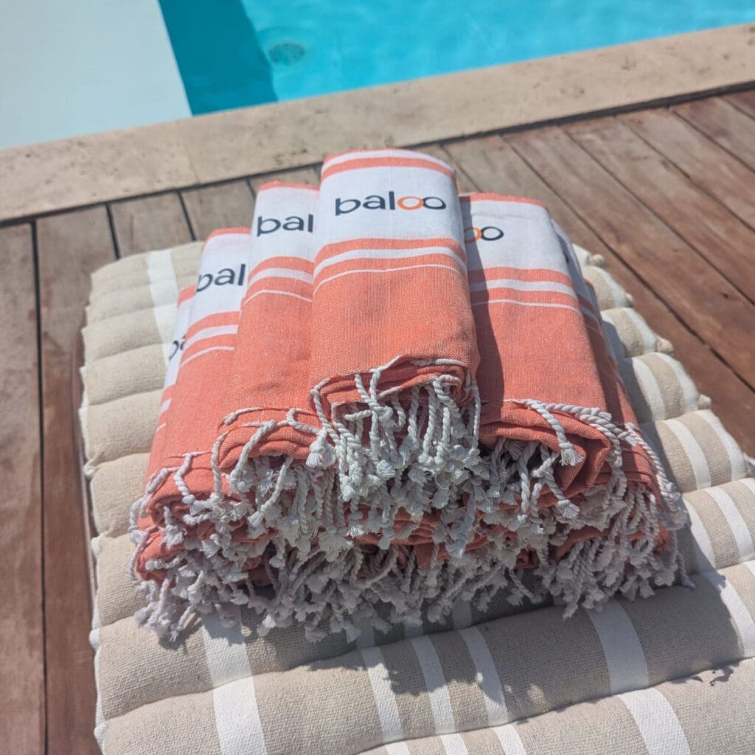 Plusieurs foutas orange pliées et empilées sur un coussin rayé beige, posées au bord d'une piscine.